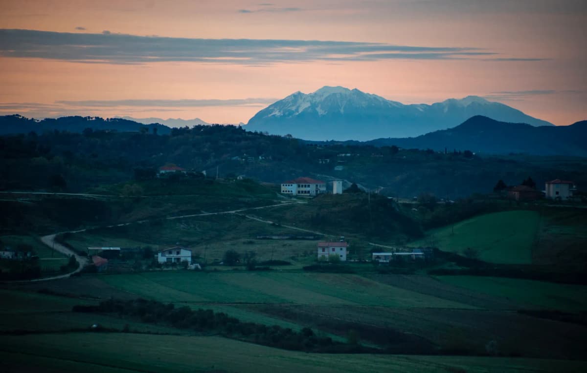 A tranquil rural scene in Albania, showcasing rolling hills and mountains under a sunset sky.